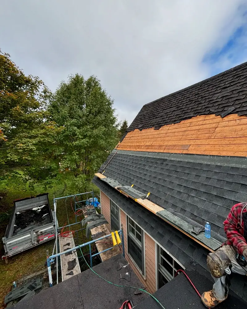 Roofer on a roof stripped back to bare plywood beside new dark shingles